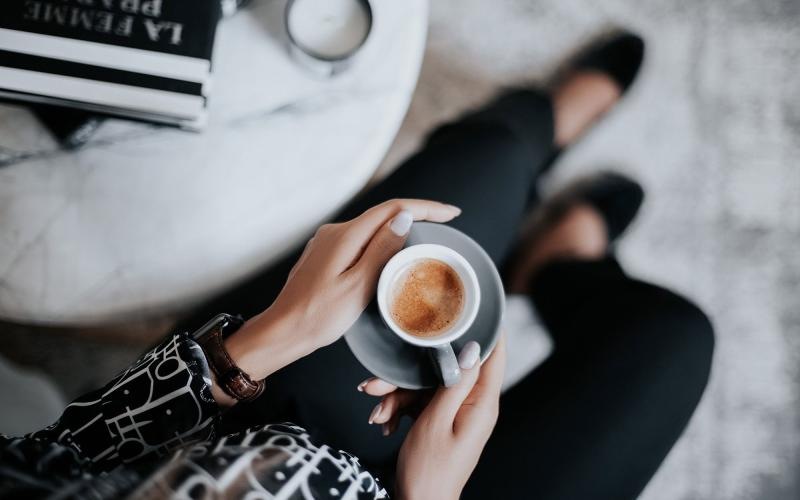 business woman sits next to a marble table with books and holds a saucer with a cup of espresso